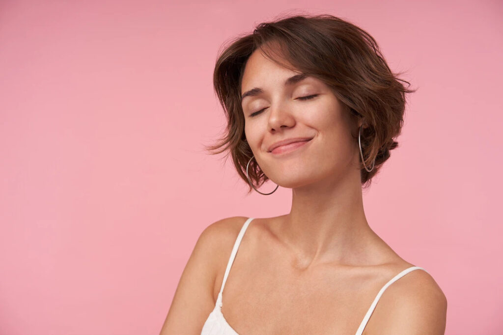 Pleasant looking young woman lady with short brown hair wearing white top while standing, keeping eyes closed and smiling gently